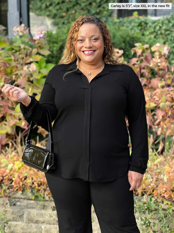 Miik model Carley (5'2", xxlarge) smiling wearing an all black outfit: Miik's Lucia collared shirt, Reed pants while holding a shoulder bag 