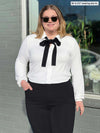 Miik model Bri (5'5", xlarge) smiling while standing in front of a window wearing Miik's Lucia collared shirt in white with a tie bow in black and a black dressy pant 