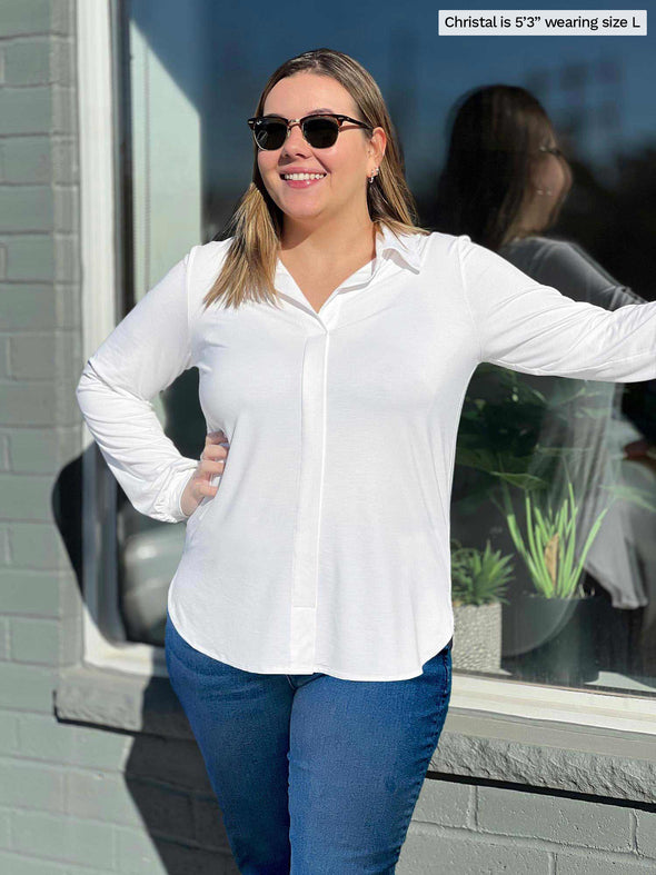 Miik model Christal (5'3", large) smiling and looking away while standing in front of a window wearing Miik's Lucia collared shirt with jeans and sunglasses 