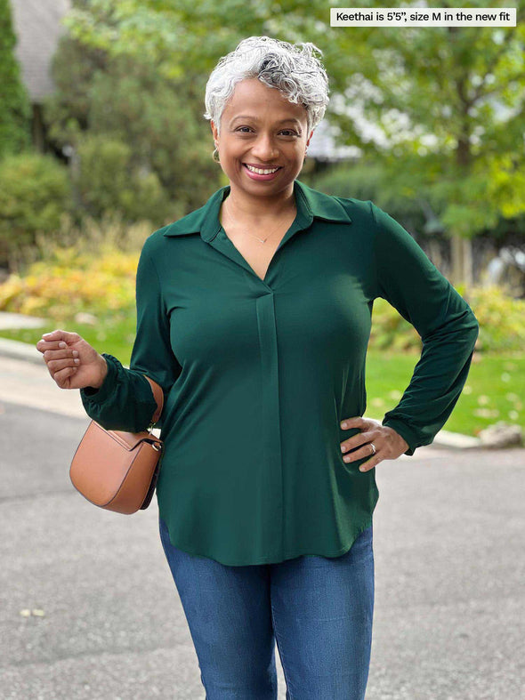 Miik model Keethai (5'5", medium) smiling wearing Miik's Lucia collared shirt in green pine in the new fit with jeans and a brown purse