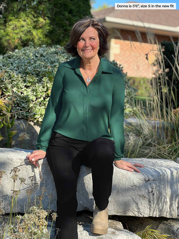 Miik founder Donna (5'6", small) sitting on a rock and looking away wearing Miik's Lucia collard shirt in green pine in the new fit along with a black pant and neutral boots