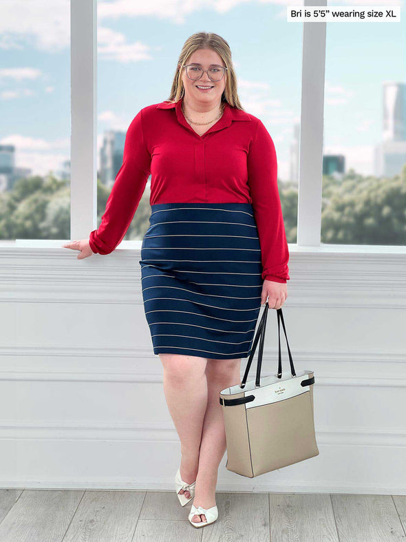 Miik model Bri (5'5", xlarge) standing in front of a window wearing a pencil striped skirt along with Miik's Lucia collared shirt in poppy red