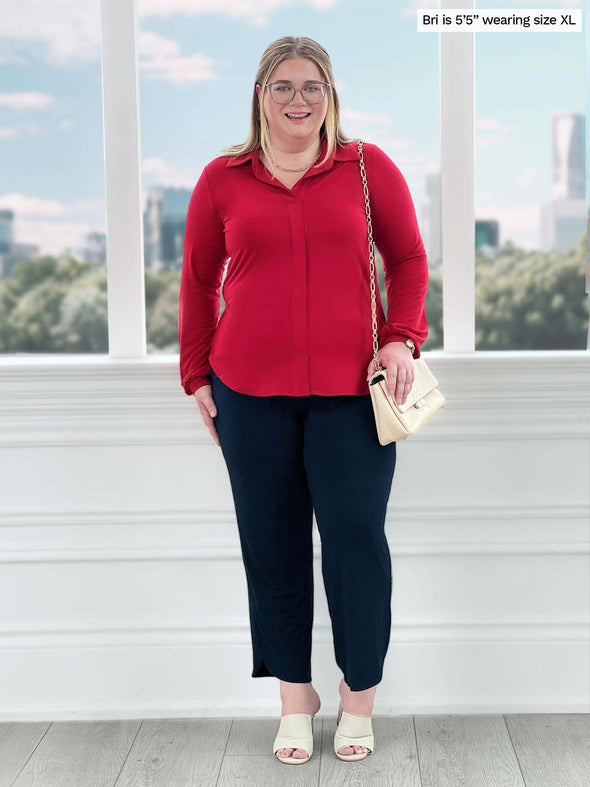 Miik model Bri (5'5", xlarge) smiling while standing in front of a window wearing Miik's Lucia collared shirt in poppy red with a navy capri pant 