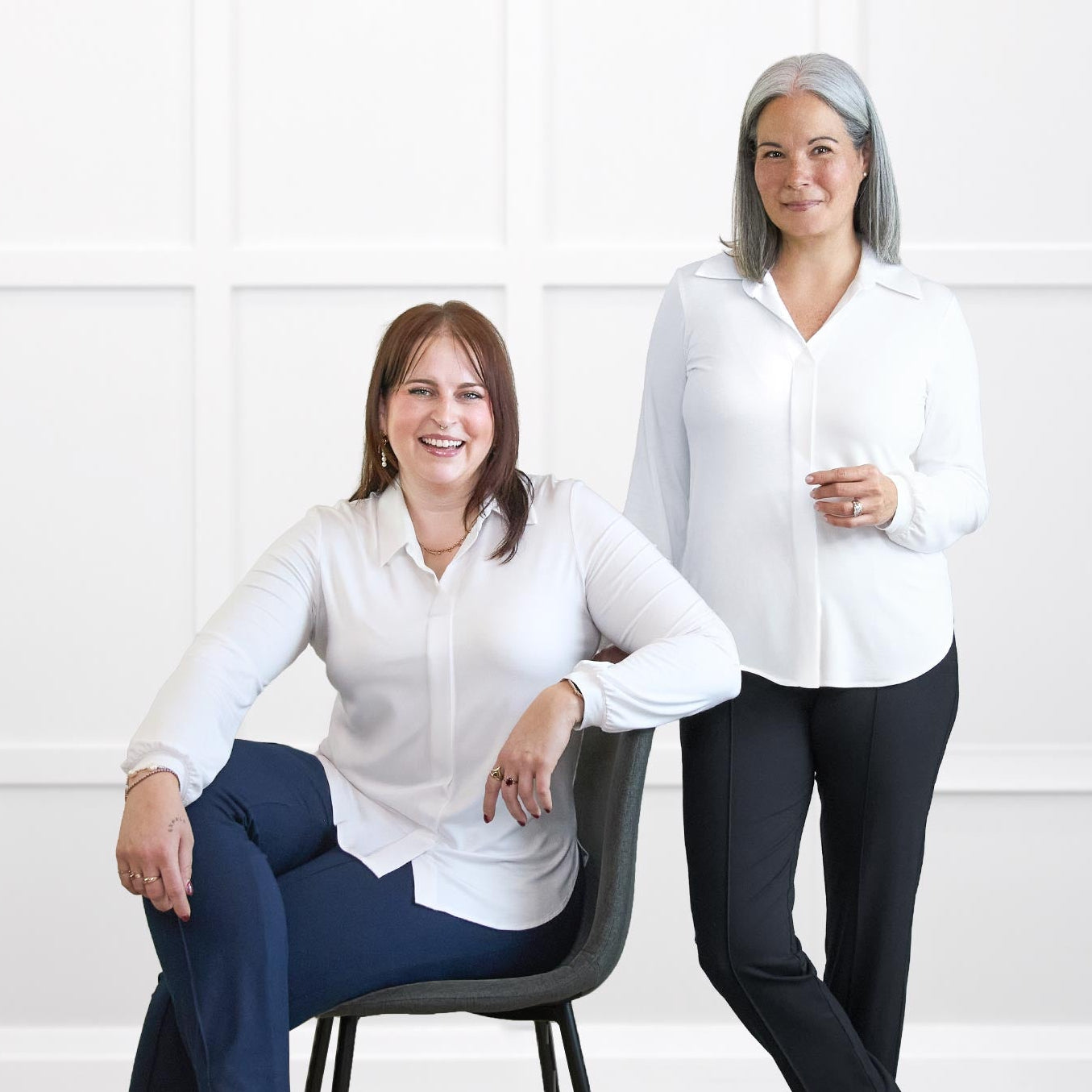 Two women in white shirts and dark pants sitting on a chair and standing against a white wall.