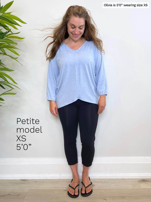 Woman standing in front of a white wall looking down wearing Miik's Lucy capri legging in navy with a light blue long sleeve top. Description of a petite model is also in the image, size XS 5"0