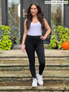 Miik model Yasmine (5'0", xsmall) wearing Miik's Lucy mid-rise legging in black with a white tank top and sneakers.