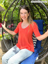 Miik model Johanna (five feet six, xsmall) sitting on a outdoor chair and smiling wearing Miik's Luisa half sleeve high low top in papaya melange with jeans 