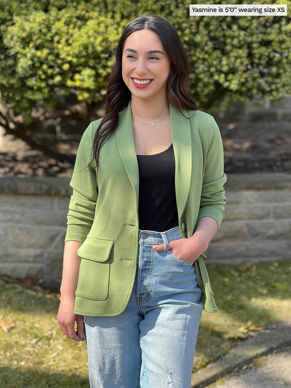 Woman standing in nature wearing Miik's Maeva LightLuxe washable blazer in moss green over a black tank and jeans.