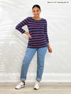 Miik model Meron (5'3", xsmall) smiling while standing in front of a white wall wearing Miik's Mahala boatneck breton top in beach house stripe with jeans and sneakers 