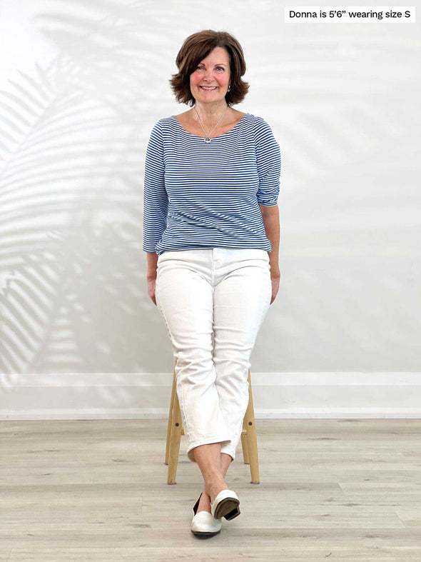 Woman sitting in front of a wall wearing Miik's Mahala boatneck breton top in blue stripe with white jeans.