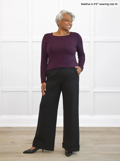 Woman wearing a purple long-sleeve top and black pants against a white background 