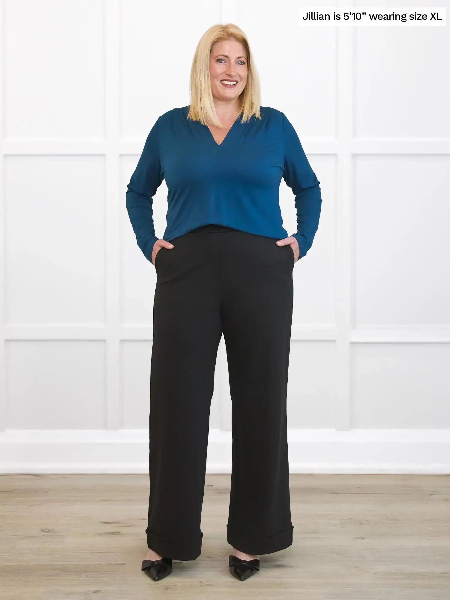 Woman wearing a blue top and black pants against a white wall. 