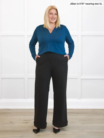 Woman wearing a blue top and black pants against a white wall. 