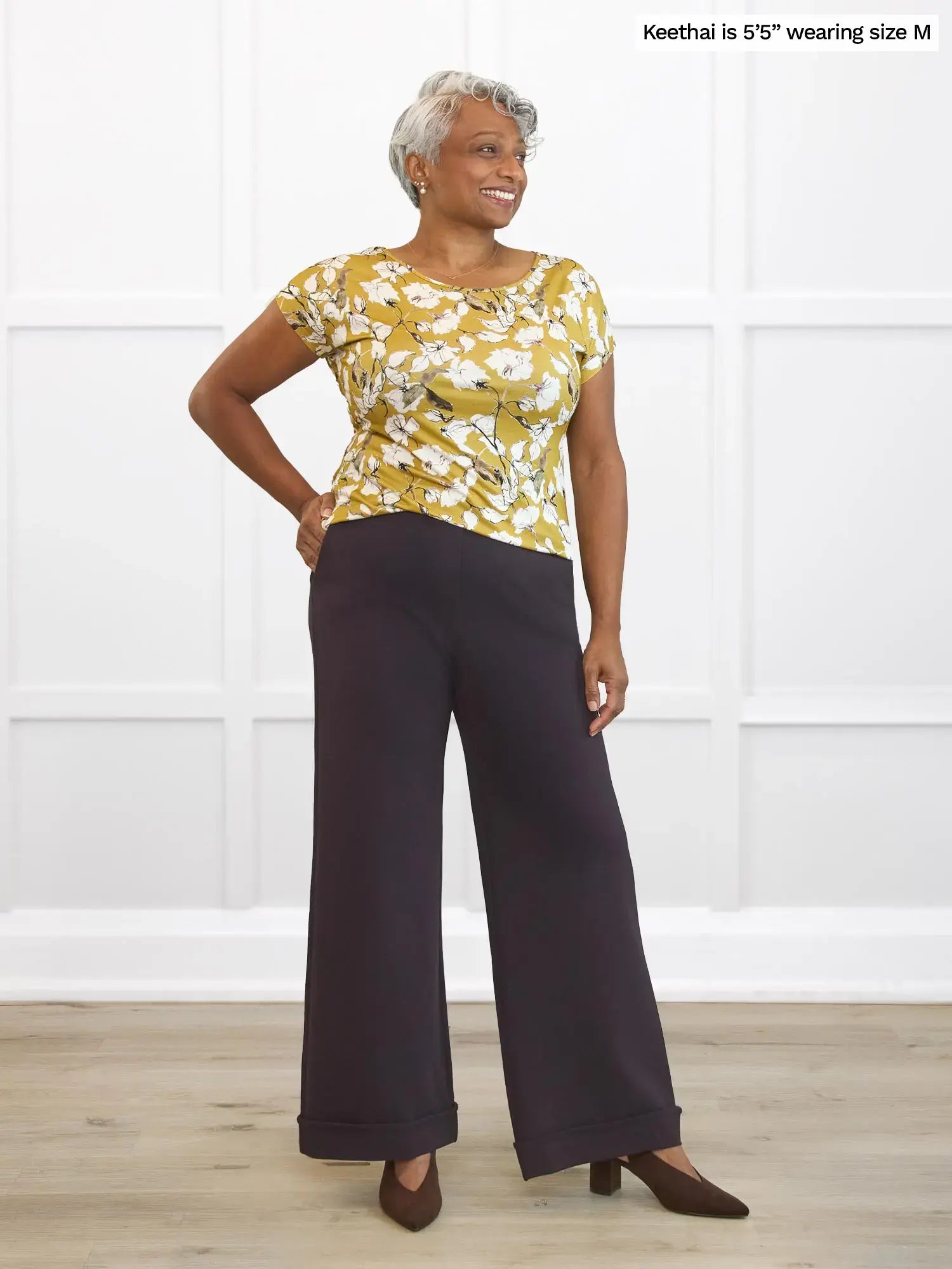 Woman wearing a yellow floral top and dark brown pants against a white background 