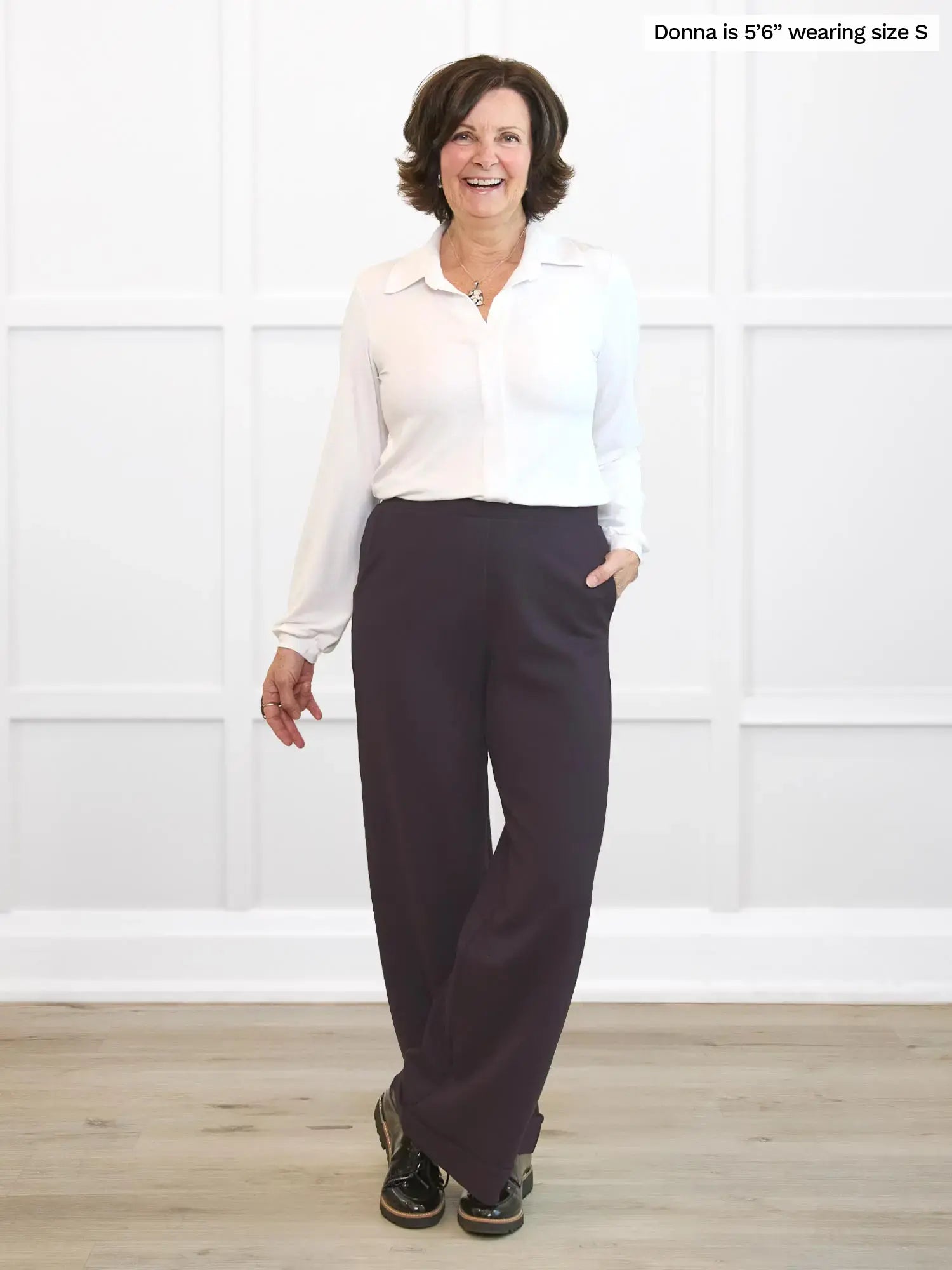 Woman wearing a white shirt and dark brown pants standing against a white wall. 