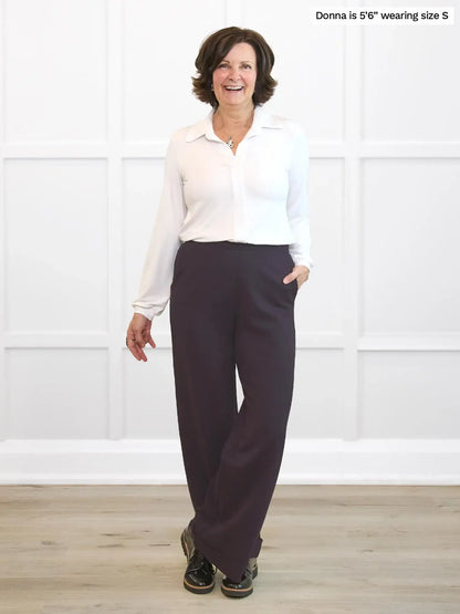 Woman wearing a white shirt and dark brown pants standing against a white wall. 