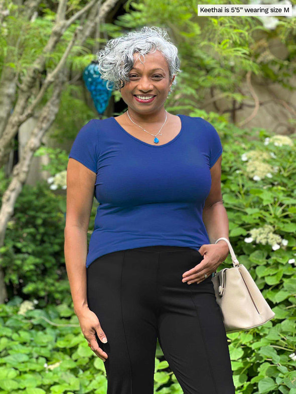Miik model Keethai (five feet five, medium) smiling while standing in a garden wearing a black pant along with Miik's Marianna reversible classic tee in blueberry and a beige purse 