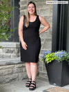 Miik model Christal (five feet three, large) standing in front of a brick wall smiling wearing Miik's Maryse reversible sweetheart dress black reversed with matching colour sandals 