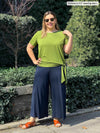 Miik model Christal (5'3", large) smiling wearing a wide leg flare pant in navy along with Miik's Melody side tie top in green moss