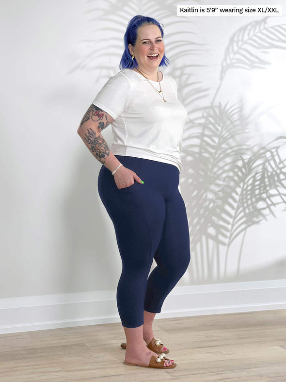 Miik model Kaitlin (5'9", xxlarge) standing sideway in front of a white wall wearing a capri navy legging along with Miik's Melody side tie top in white 