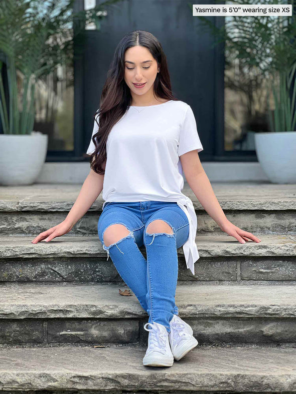 Miik model Yasmine (5'0, xsmall, petite) sitting on a stairway looking down wearing Miik's Melody side tie top in white with a ripped jeans 