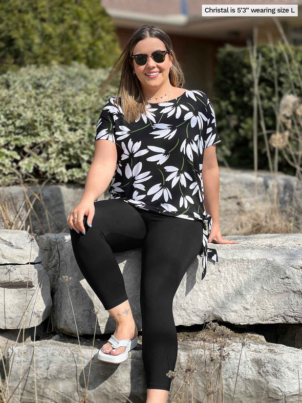 Miik model Christal (5'3", large) sitting on a rock smiling while wearing Miik's Melody side tie top in white lily and capri black legging 