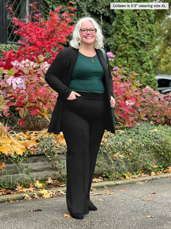 Miik model Colleen (5'3", xlarge) standing in front of a garden smiling wearing a black pant, green pine top along with Miik's Montana fleece pocket cardigan in black