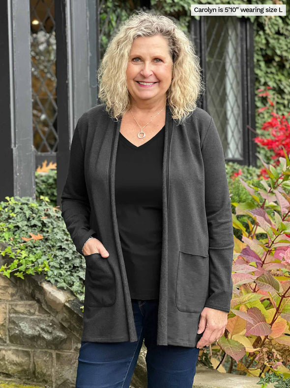 Miik model Carolyn (5'10", large) smiling with hand on the pocket wearing Miik's Montana fleece pocket cardigan in charcoal along with a black v-neck tee and jeans