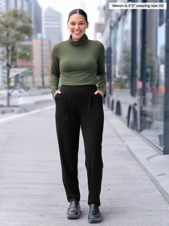 Miik model Meron (5'3", xsmall) smiling with hands on pockets wearing Miik's Nala pleated tapered pant in black with a turtleneck top in sage melange  