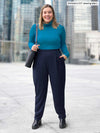 Miik model Christal (5'3", large) smiling with one hand on the pocket wearing Miik's Nala pleated tapered pant in navy and a turtleneck top in teal melange, black boots