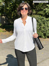 Woman standing outside wearing Miik's Neruda band collar long sleeve shirt in white with grey pants.