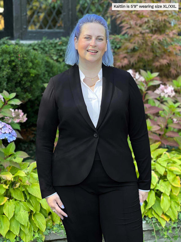 Woman standing in front of plants wearing Miik's Neruda band collar long sleeve shirt in white under a black blazer with black pants.