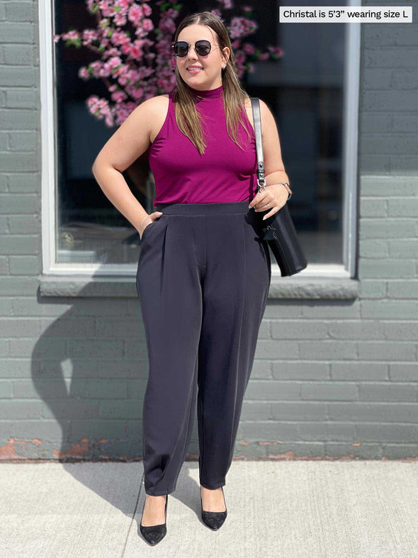 Miik model Christal (five feet three, size large) standing in front of a window/brick wall wearing Miik's Nida pleat front tapered pant in graphite along with a ruby mock neck tank and a shoulder bag