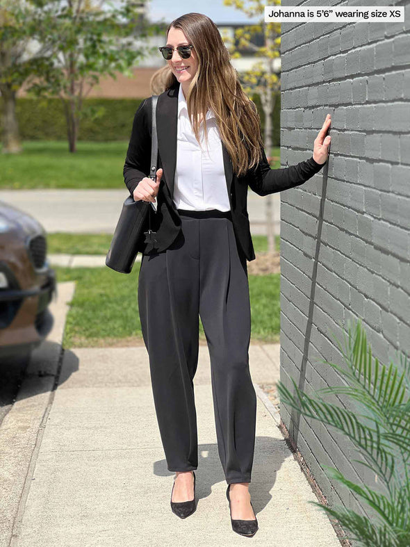 Miik model Johanna (five feet six, size xsmall) standing next to a brick wall wearing Miik's Nida pleat front tapered pant in graphite along with a white collared shirt and a black blazer