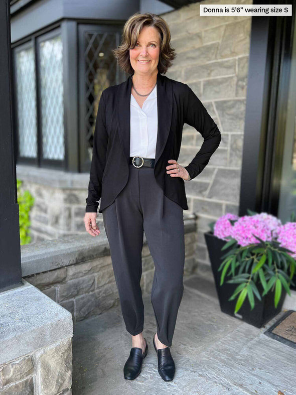 Miik founder Donna (five feet six, small) smiling wearing Miik's Nida pleat front tapered pant in graphite with a white collared shirt and a black blazer/cardigan 