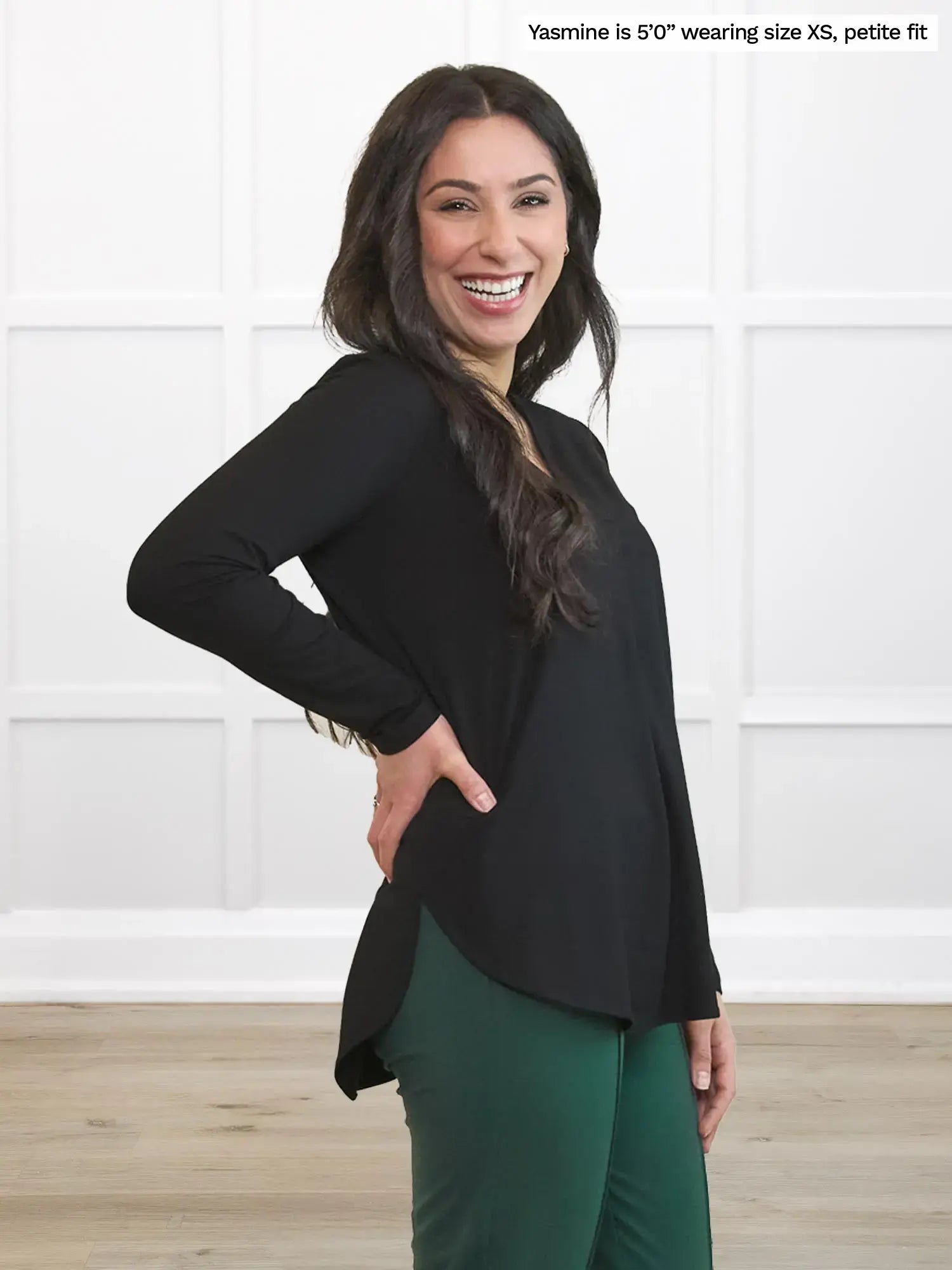 Woman wearing a black long-sleeve top and green pants against a white paneled wall. 