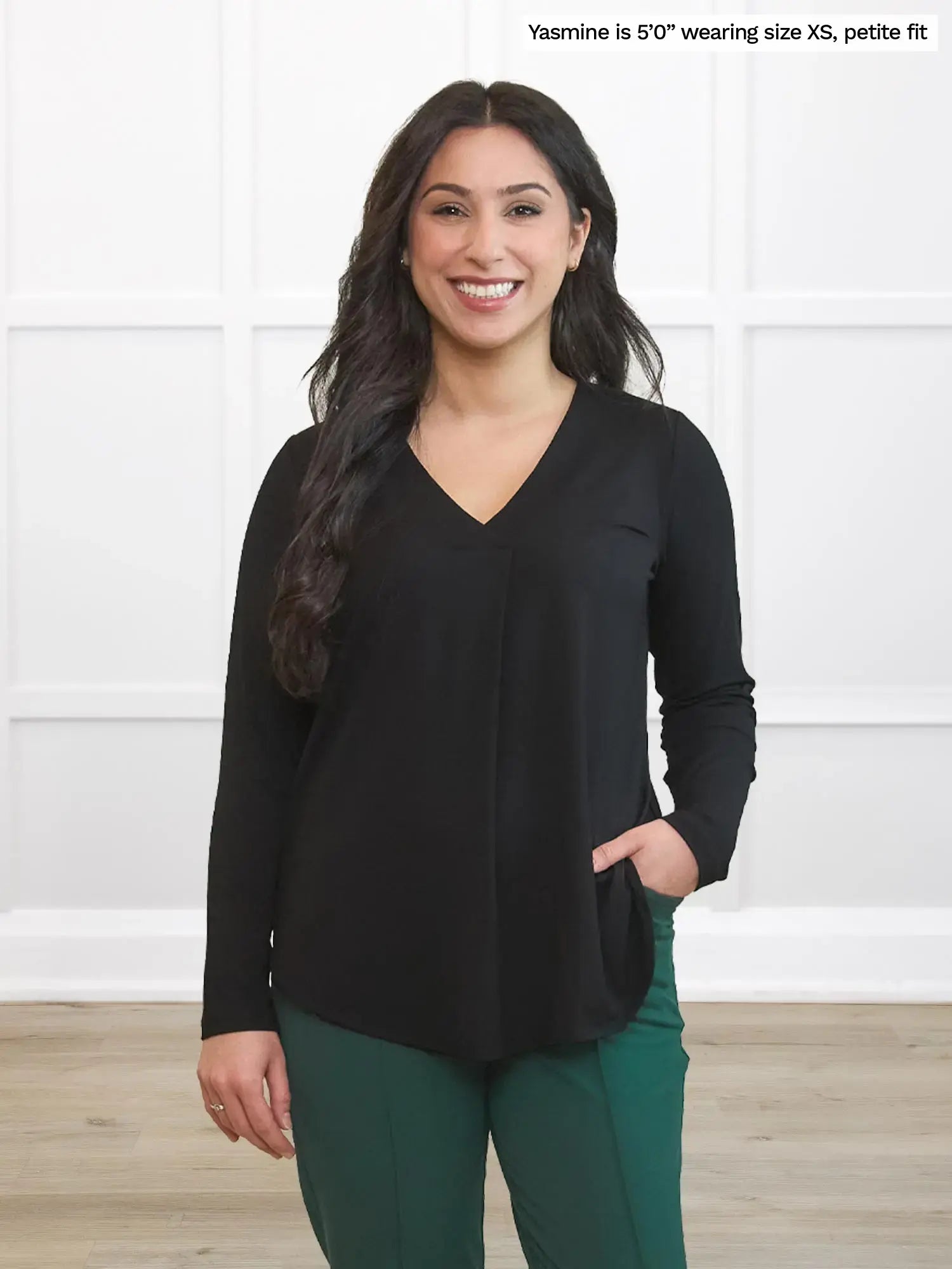 Woman wearing a black long-sleeve top and green pants against a white wall. 