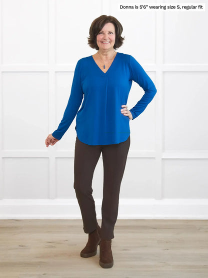 Woman wearing a blue long sleeve top and brown pants standing against a white wall. 
