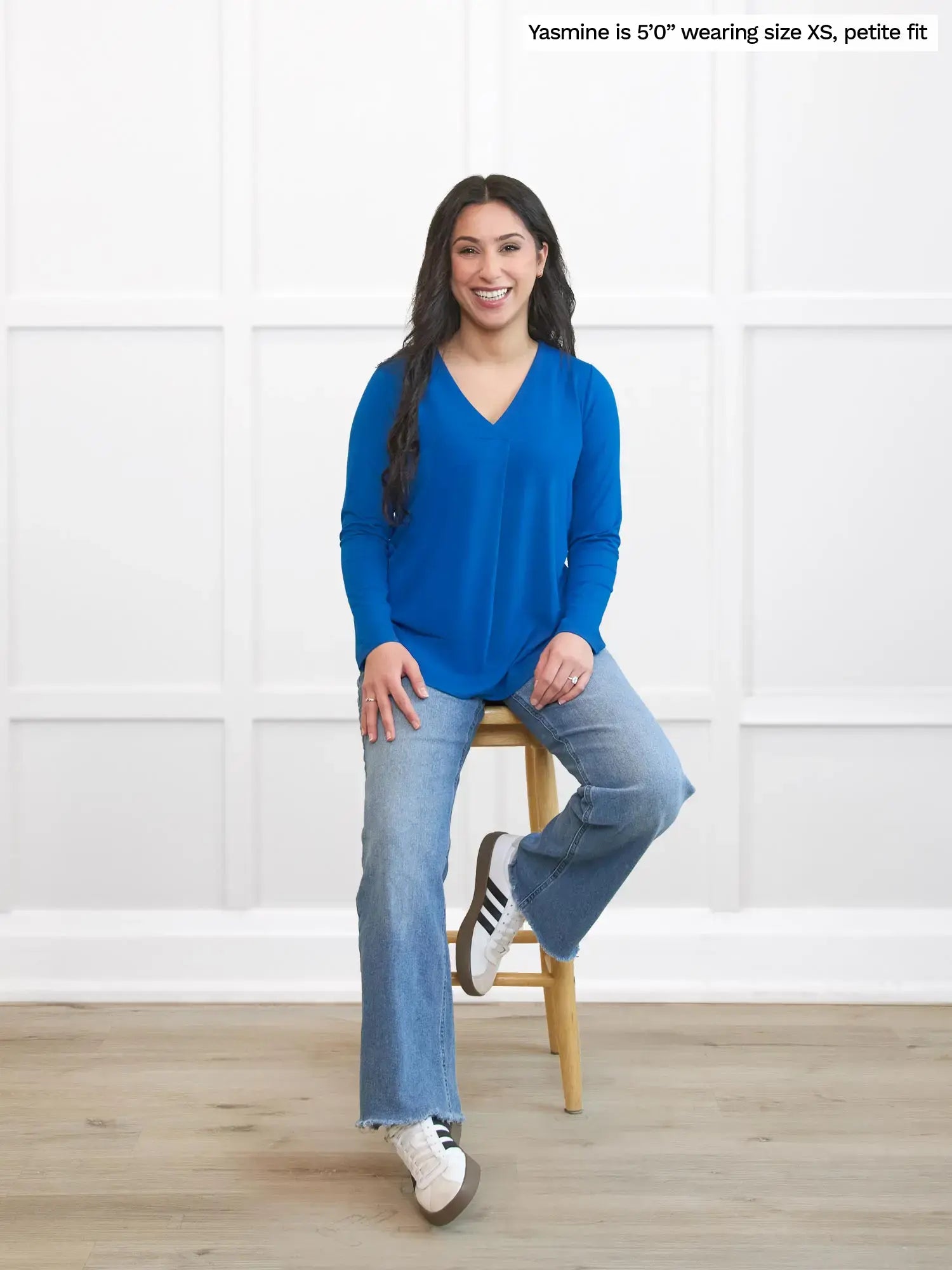 Woman wearing a blue top and jeans sitting on a stool against a white wall. 