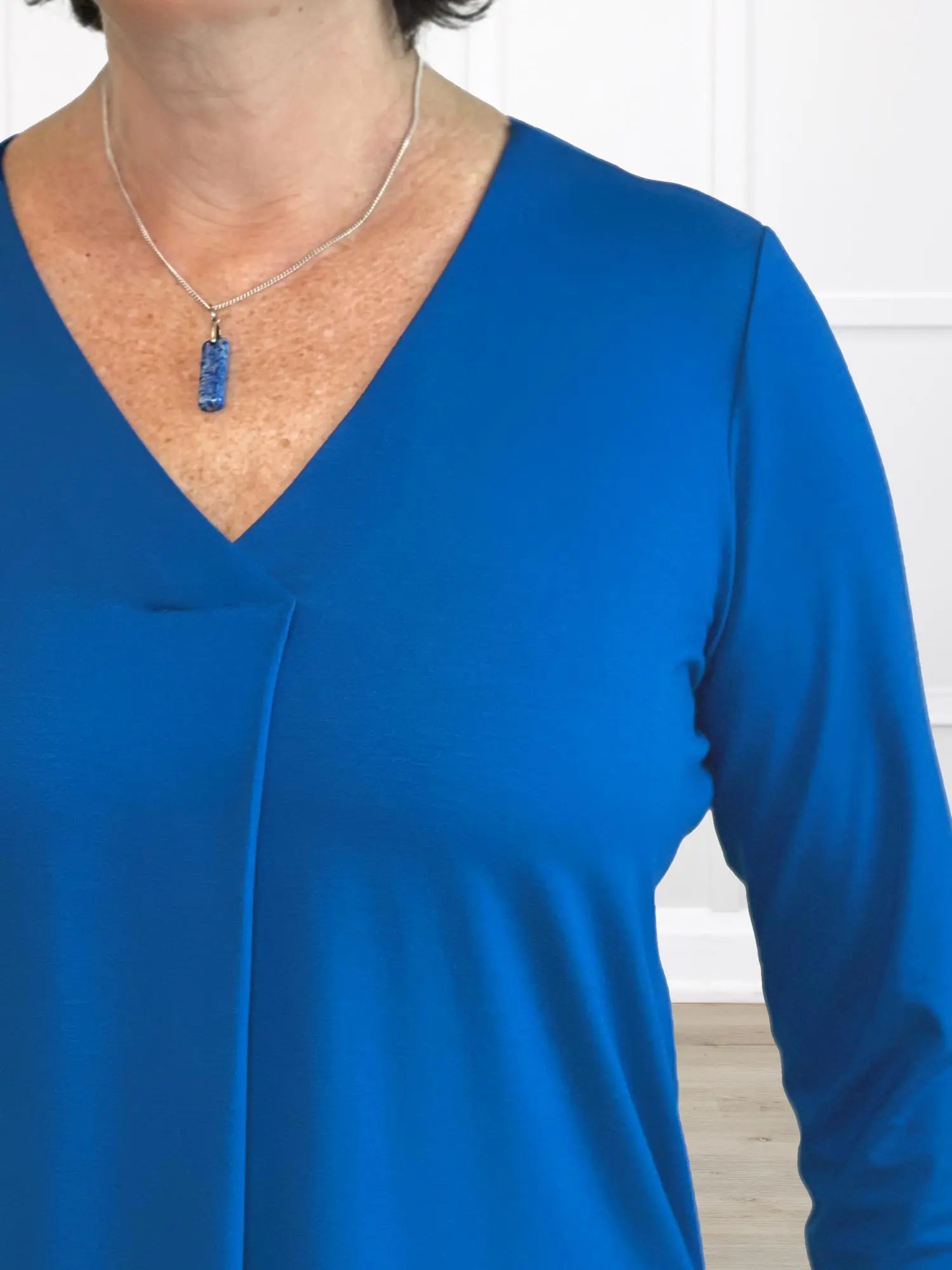 Person wearing a blue long-sleeve top with a necklace against a white wall. 
