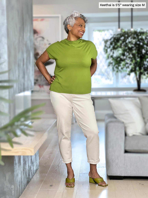 Miik model Keethai (5'5", medium) smiling while standing in a living room wearing Miik's Noah draped dolman top in green moss and a white jeans 