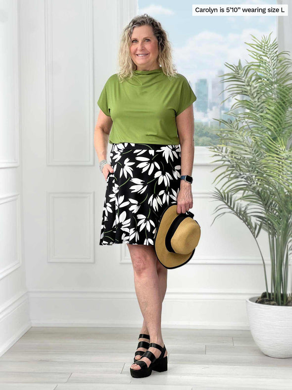 Miik model Carolyn (5'10", large) smiling while standing in front of a white wall wearing a printed skirt with Miik's Noah draped dolman top in green moss