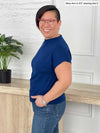 Miik model Mary-Ann (5'0", small) smiling standing sideway wearing Miik's Noah draped dolman top in ink blue with jeans 