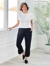 Miik founder Donna (5'6", small) smiling while standing next to a window/white wall wearing Miik's Noah draped dolman top in white tucked in a capri pant in black