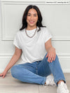 Miik model Yasmine (5'0", xsmall, petite) sitting on the floor and smiling wearing Miik's Noah draped dolman top in white with jeans and sneakers 