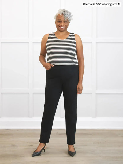 Woman wearing a striped sleeveless top and black pants standing against a white wall. 