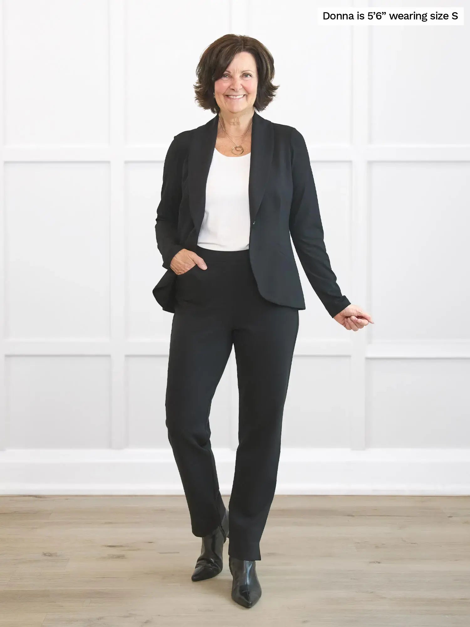 Woman wearing a black suit standing in a room with white walls and wooden floor. 