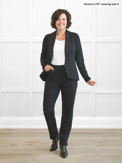 Woman wearing a black suit standing in a room with white walls and wooden floor. 