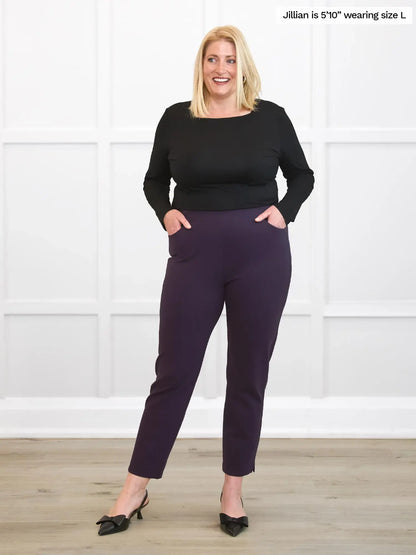 Woman wearing a black top and purple pants standing against a white wall. 