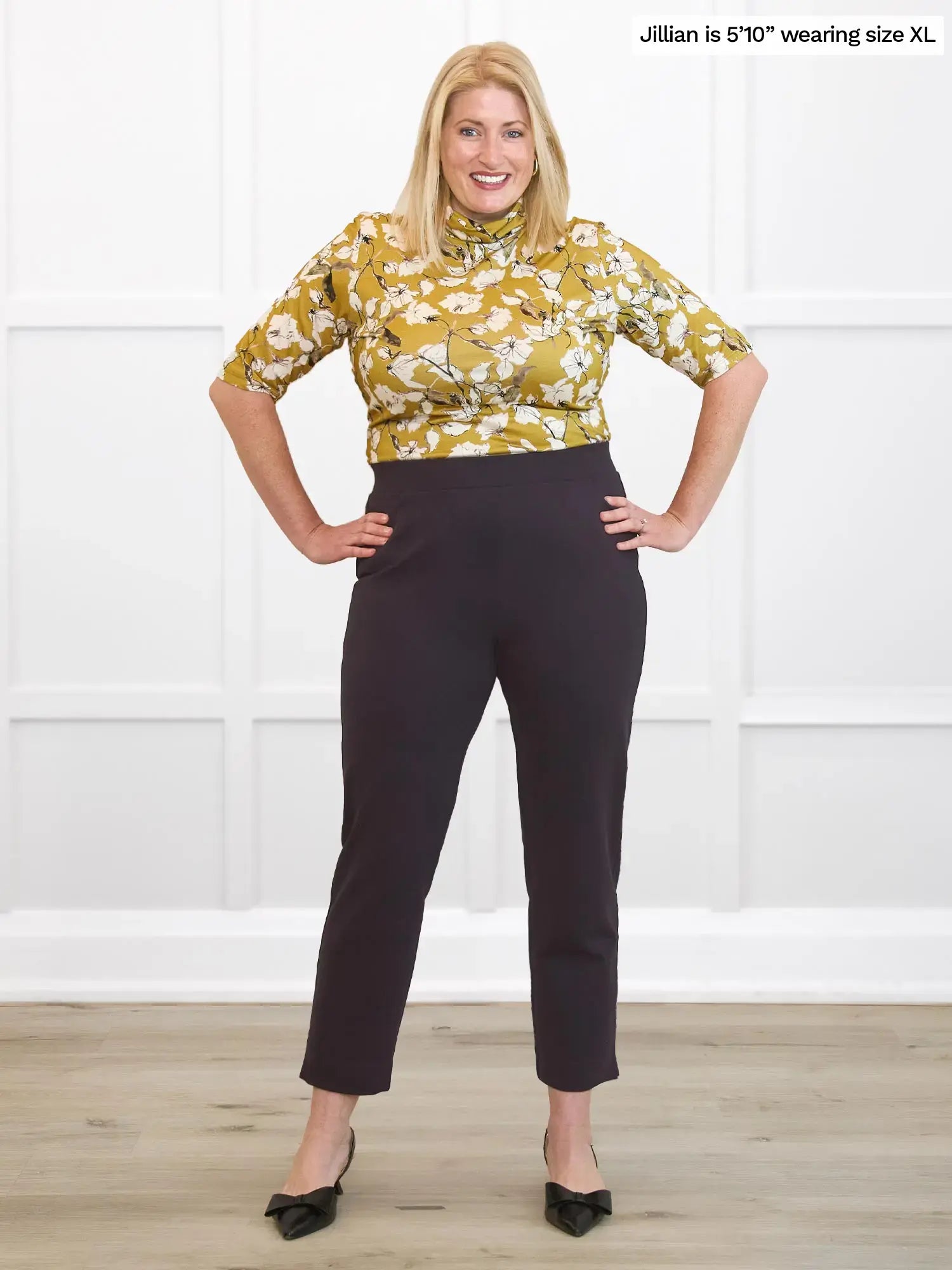 Woman wearing a yellow floral blouse and dark brown pants against a white background 